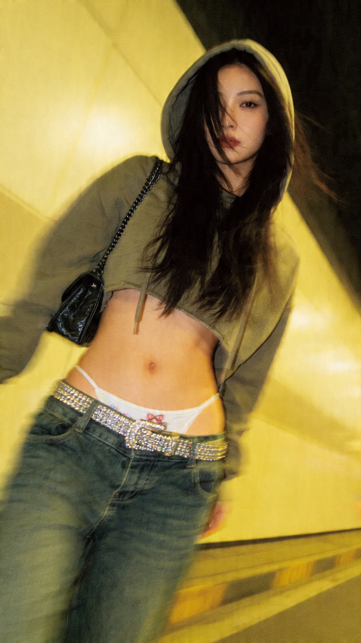 a lo-fi 2015s-era, diagonally taken, low-quality iPhone photo of a 20-year-old baddie. The shot is imperfect — grainy, off-center, motion-blurred, and raw. The lighting is uneven and yellowish, like a flash bounced off a mirror or cheap LED strip. She’s captured head-to-hip, wearing a cropped hoodie and low-rise jeans with a rhinestone belt and playfull tanga peeking, a tiny purse hanging off her shoulder. Her long hair partially covers her face as she moves mid-pose, expression unreadable.
